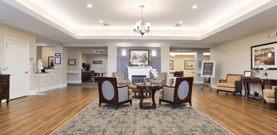 A spacious and well-lit senior living facility common area with a seating arrangement of four cushioned chairs around a small round table on a patterned rug. The room features a fireplace with decorative items on the mantle, a chandelier, wooden flooring, and additional seating and tables along the walls. The walls are painted in neutral tones with framed artwork and a welcoming sign on an easel.