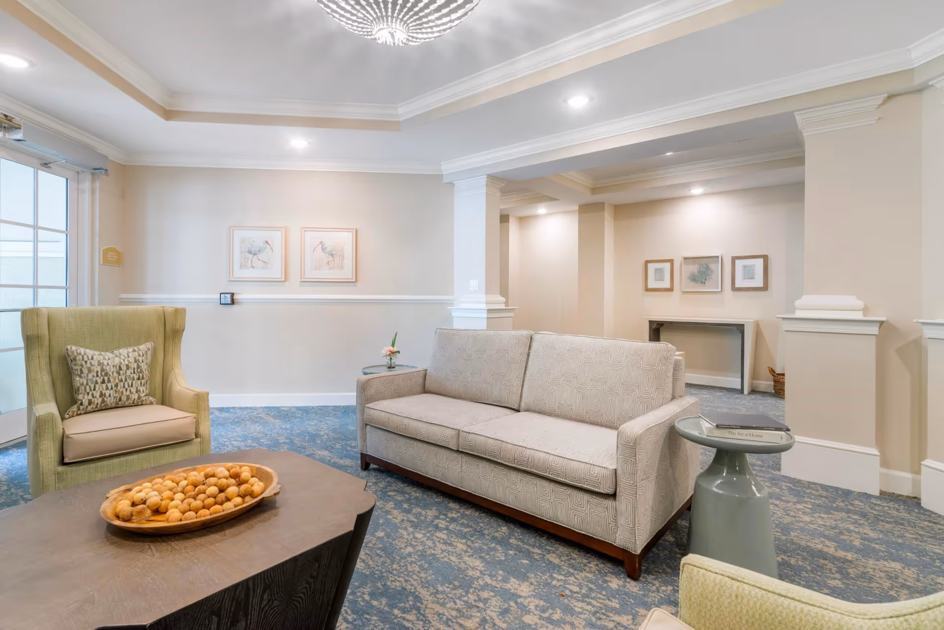 A bright seating area with a sofa, upholstered armchairs, a coffee table holding a bowl of decorative balls, and framed wall art.