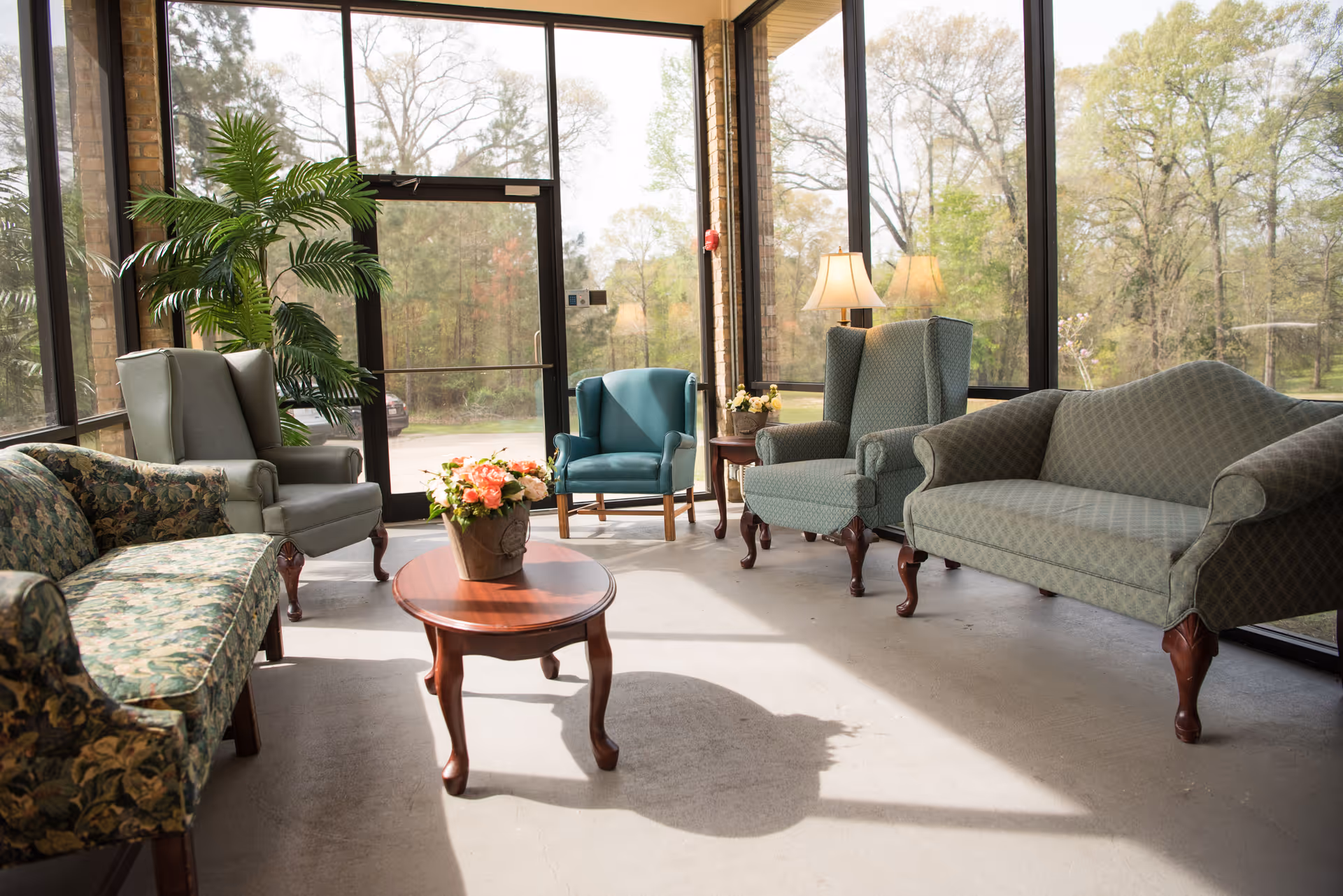 A sunlit sitting area with large floor-to-ceiling windows showing trees outside. The room contains a floral patterned sofa, two green armchairs, a blue armchair, a green loveseat, a wooden coffee table with a flower arrangement, a side table with a lamp, and a large potted plant near the glass door.