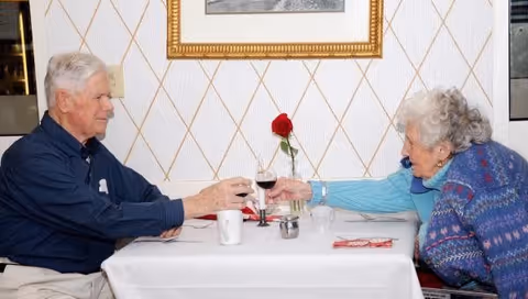 An elderly man and woman sit across a small dining table clinking wine glasses over a white tablecloth with a single red rose between them.