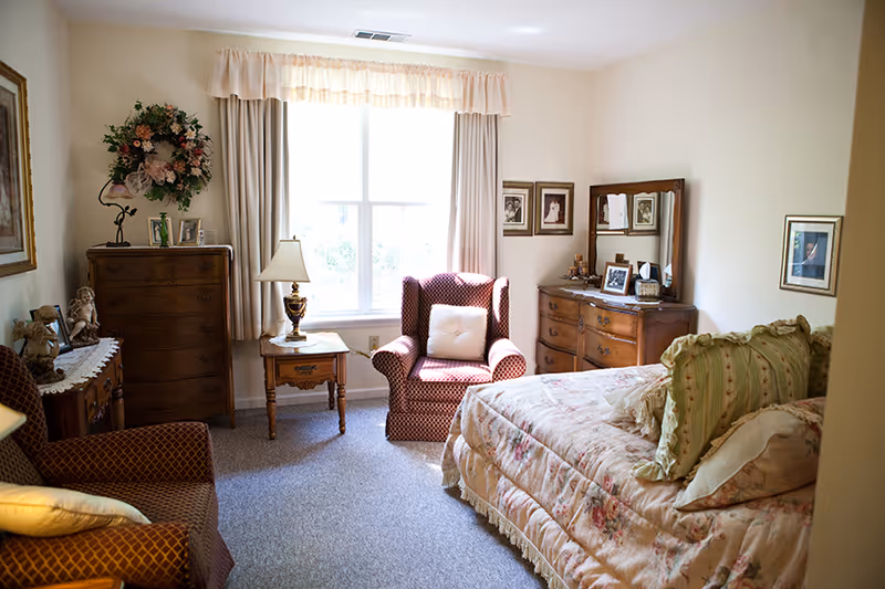 A cozy bedroom with a floral-patterned bedspread and multiple pillows, two upholstered armchairs with patterned fabric, a wooden dresser with a mirror, a tall chest of drawers, a side table with a lamp, framed pictures on the walls, and a large window with curtains letting in natural light.