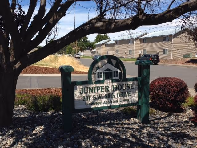 A wooden Juniper House assisted living sign sits in landscaped gravel and shrubs with the facility buildings and a street behind it.
