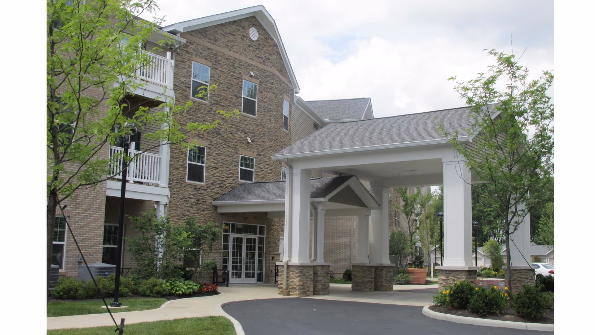 Exterior view of a multi-story senior living facility building with stone and brick facade, a covered entrance supported by white columns with stone bases, surrounded by landscaped greenery and trees under a cloudy sky.