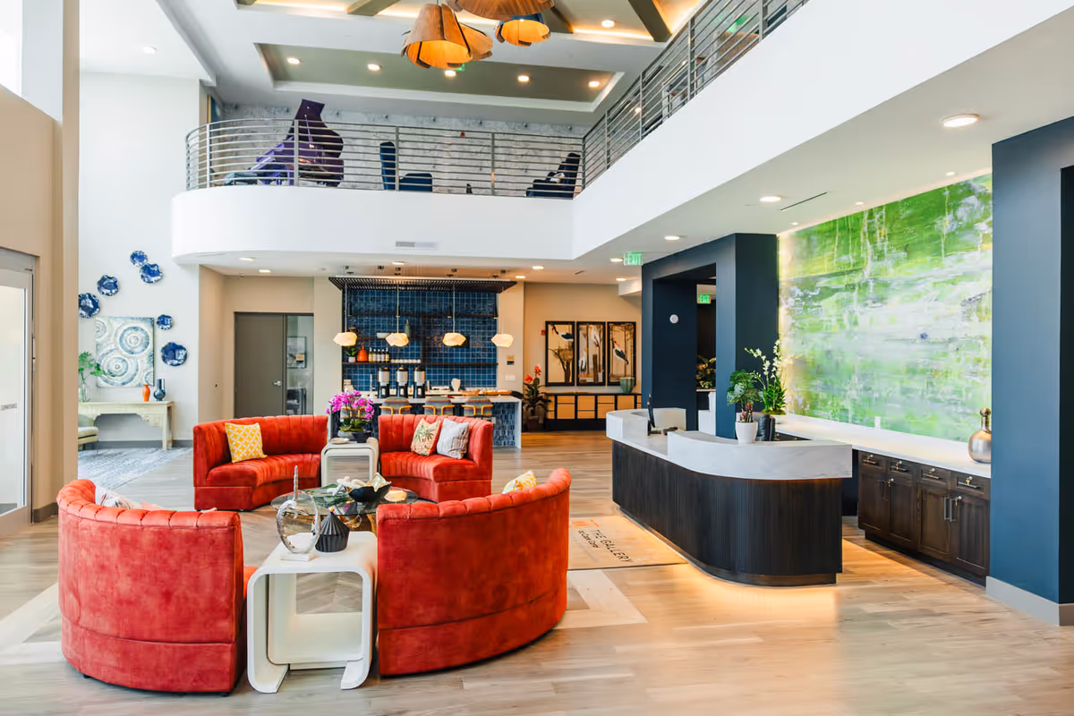 Bright two-story senior living facility lobby with curved red sofas, a reception desk, and a seating area under a mezzanine.