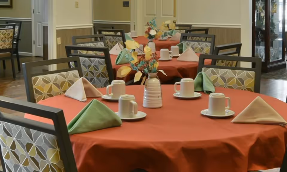 Dining room with round tables covered in red tablecloths, each set with white cups and saucers, folded green and beige napkins, and a vase with artificial flowers as a centerpiece. Chairs with patterned upholstery surround the tables.