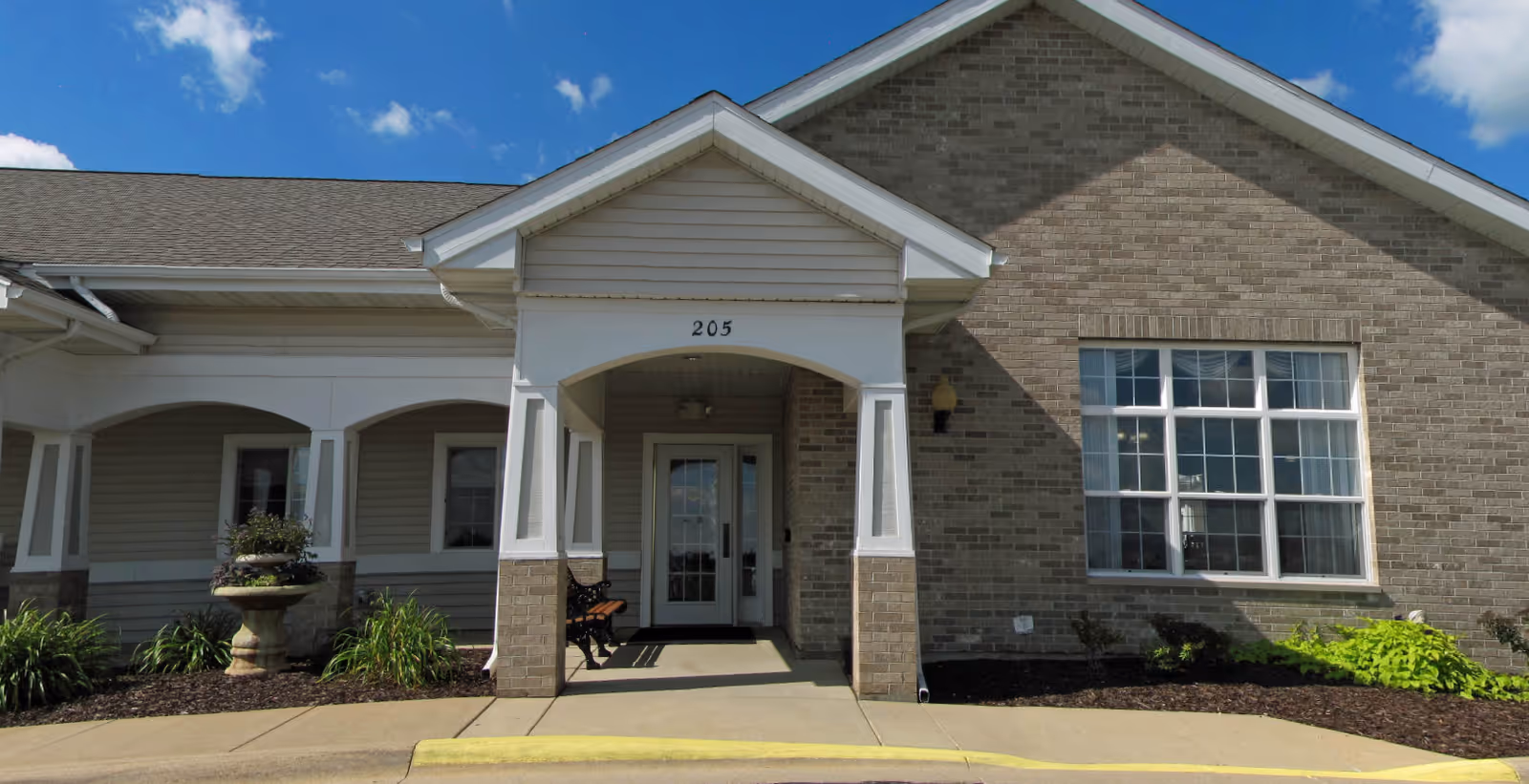 Front entrance of a single-story brick building with a covered portico marked 205, a bench, and landscaped beds.