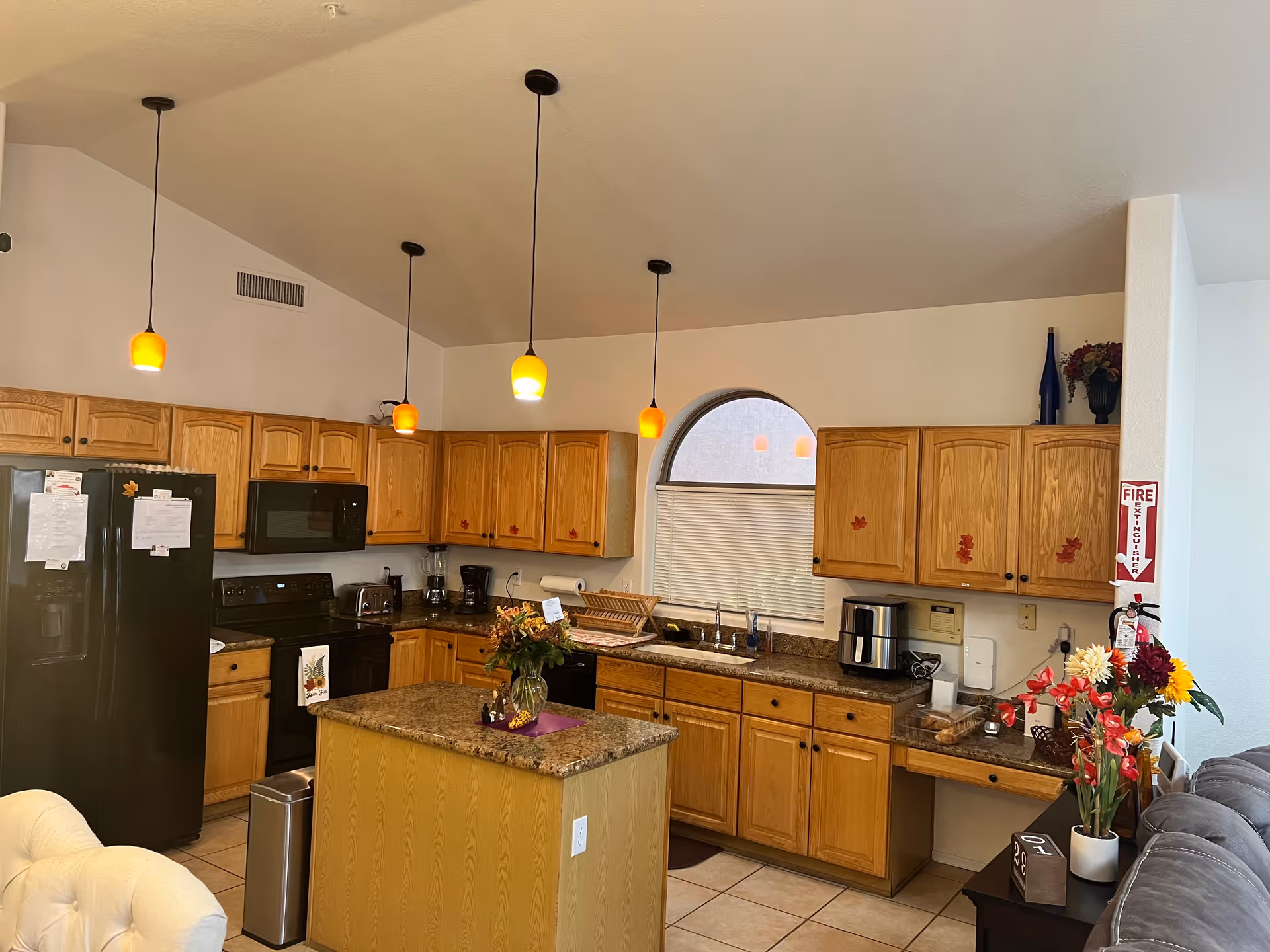 A spacious kitchen with wooden cabinets, a black refrigerator, black microwave, black stove, and granite countertops. There is a kitchen island with a flower vase on it, three hanging pendant lights with orange shades, and a window with a half-circle top and blinds. Various kitchen appliances and decorations are visible, including a fire extinguisher mounted on the wall and a bouquet of flowers on a side table next to a gray couch.