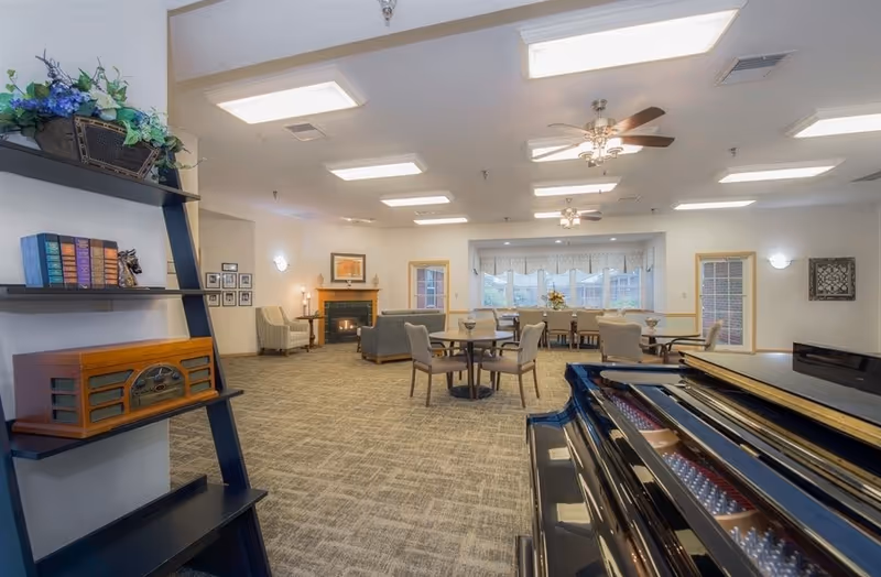 Spacious senior living common area with carpeted floor, multiple round tables with chairs, a grand piano in the foreground, a fireplace with framed artwork above it, and large windows letting in natural light. Shelves with books and decorative items are visible on the left side.