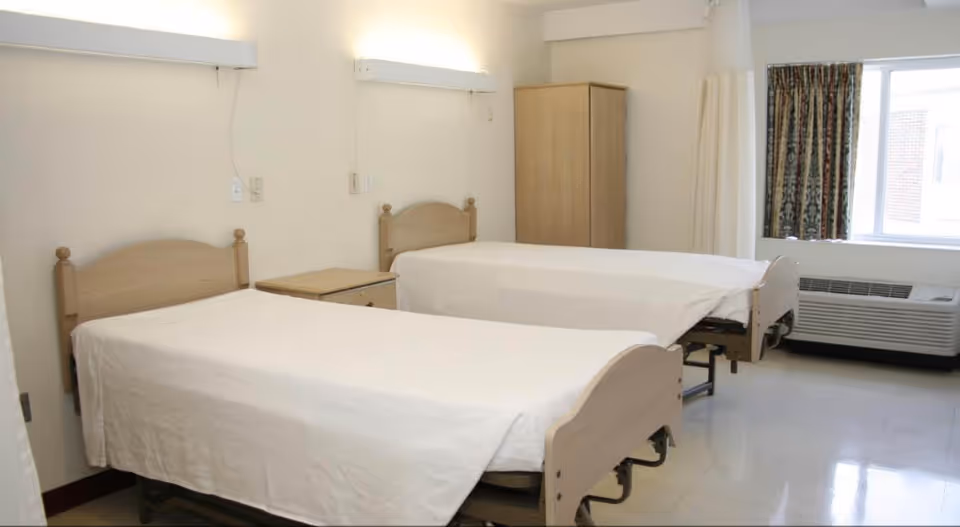A shared nursing facility bedroom with two hospital-style beds, a nightstand, a wardrobe, a window and an air conditioning unit.