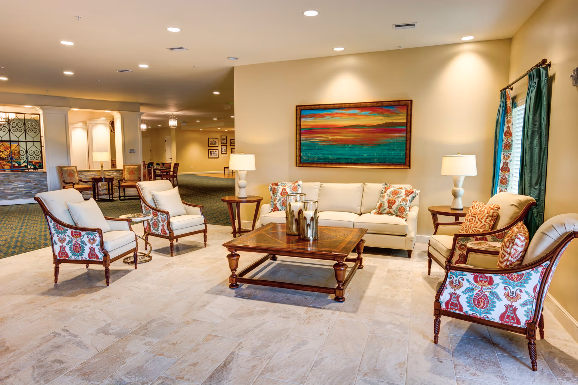 Bright furnished lounge with a sofa, patterned armchairs, wooden coffee table, lamps, and a colorful painting on the wall.