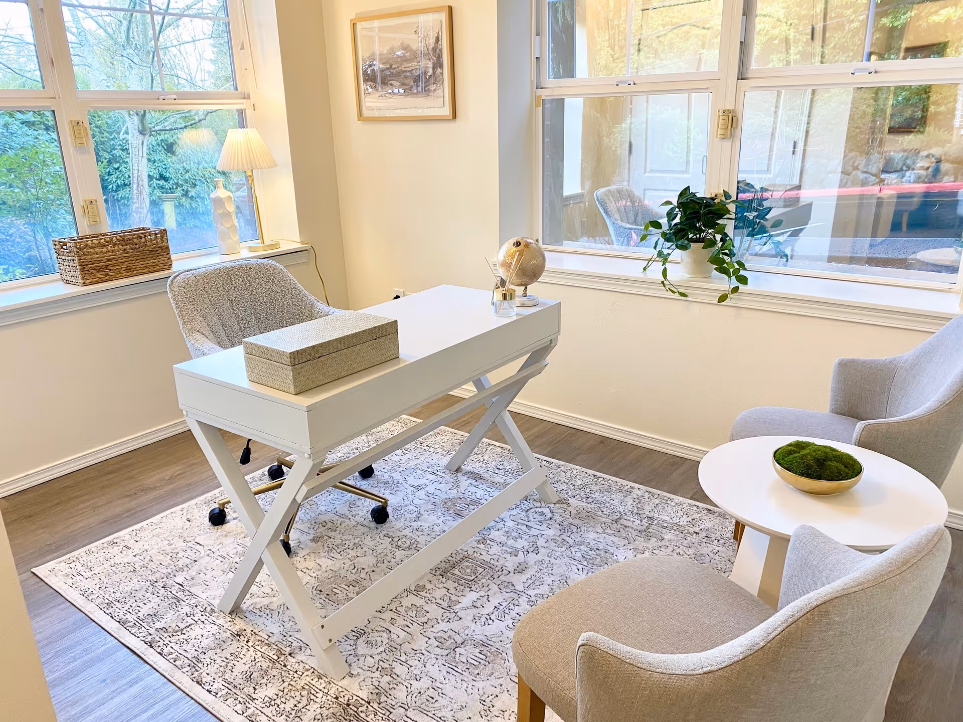A bright and cozy office space with a white desk and a cushioned swivel chair on a patterned rug. Two beige upholstered chairs are positioned around a small white round table with a decorative bowl of green moss. Large windows let in natural light and show greenery outside. A table lamp, a basket, a small globe, and a plant add decorative touches.