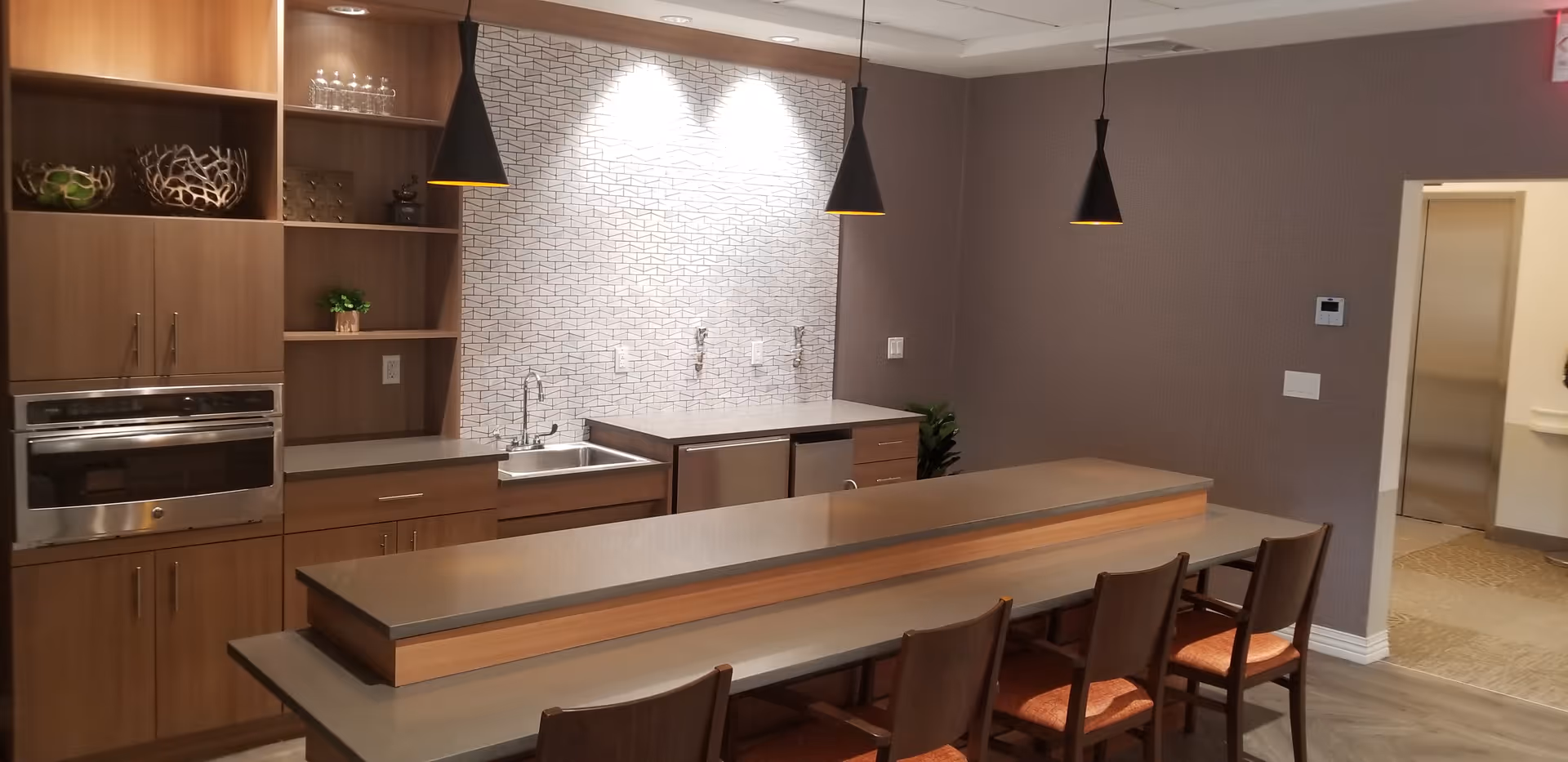 Communal kitchen and bar area with pendant lights, sink, oven, and a long counter lined with chairs.