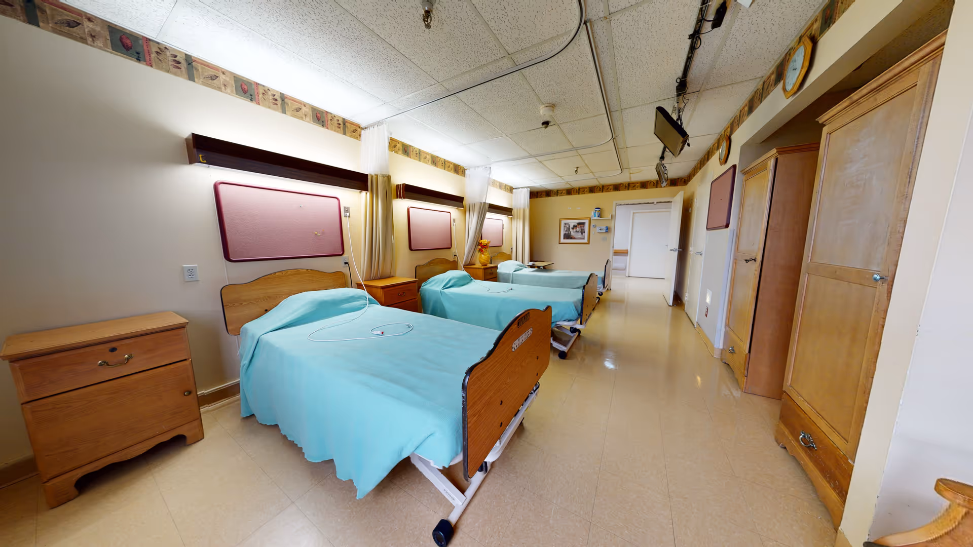 A room with two hospital-style beds covered with light blue sheets, each with a wooden headboard and footboard. There are wooden nightstands beside each bed, a wooden wardrobe, and a chair. The room has a tiled floor, a drop ceiling with fluorescent lights, and a patterned border near the ceiling. The walls are light-colored, and there are curtains between the beds for privacy.