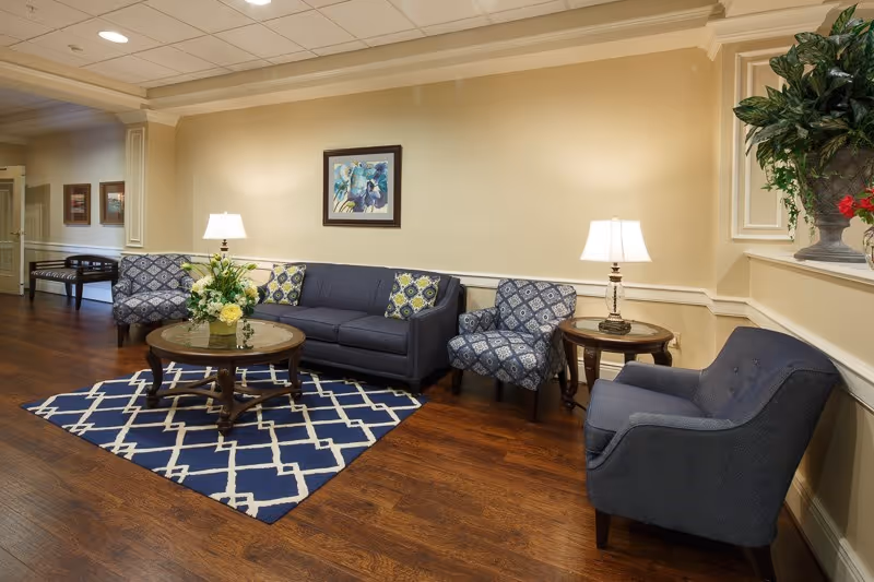 A cozy sitting area with a dark blue sofa, two patterned armchairs, and a solid blue armchair arranged around a wooden coffee table with a floral centerpiece. The floor has a blue and white geometric patterned rug. Two table lamps on side tables provide warm lighting, and a framed floral painting hangs on the beige wall behind the sofa. There is a large decorative plant on a ledge to the right.