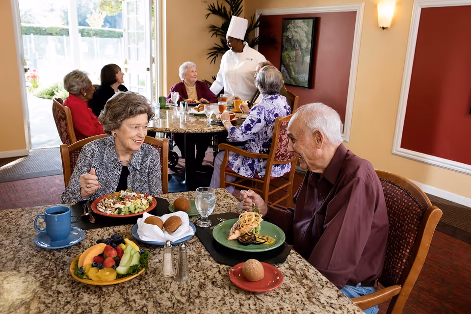 Two elderly people, a woman and a man, are sitting at a dining table enjoying a meal together in a senior living facility dining room. The woman has a salad and a cup of coffee, while the man has a plate with a main dish and a bread roll. In the background, other elderly residents are seated at another table, and a chef in a white uniform is interacting with them. The room has warm-colored walls and large windows letting in natural light.