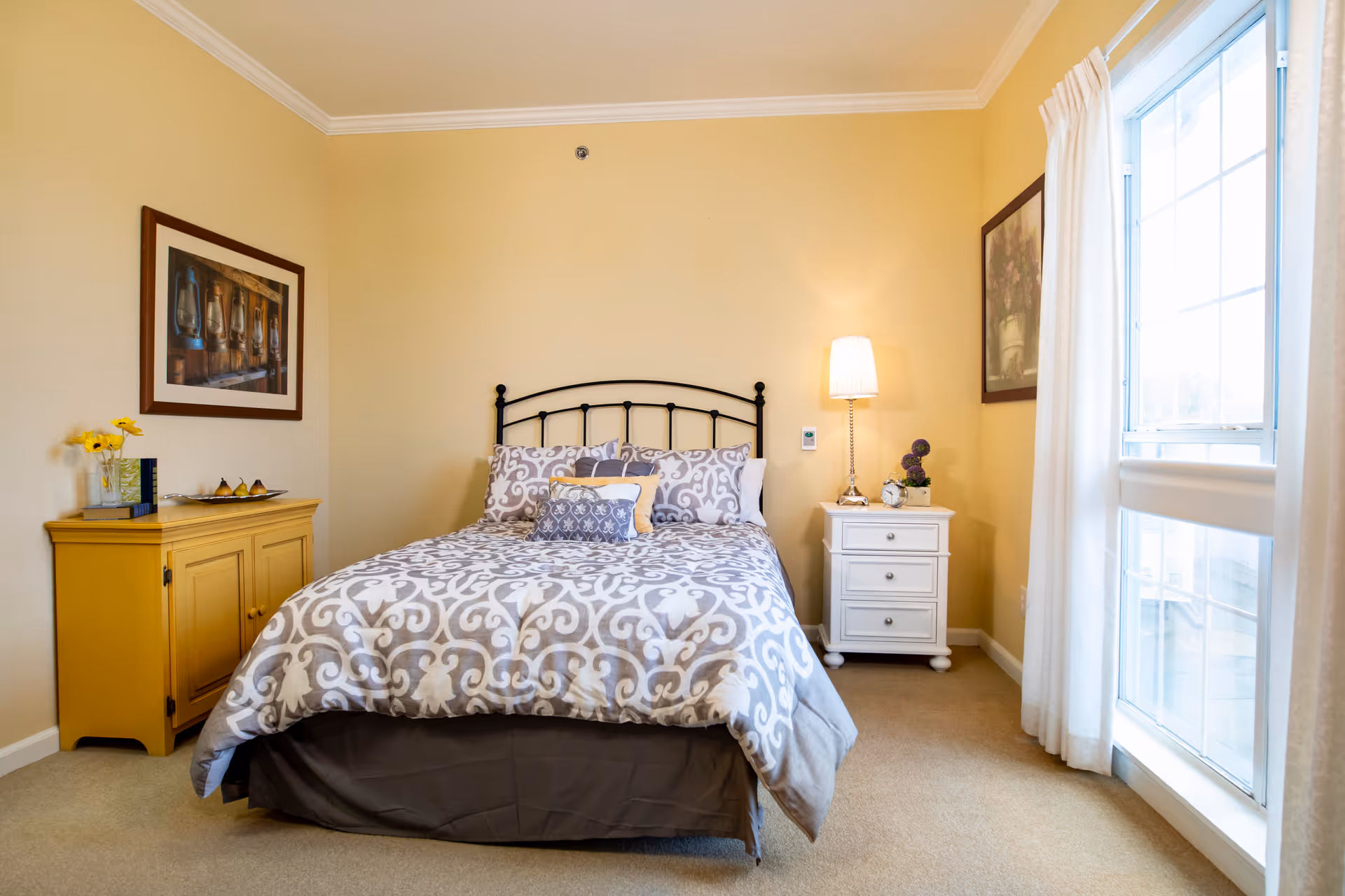 A cozy bedroom with a bed featuring a patterned gray and white comforter and multiple pillows. On the left side of the bed is a yellow cabinet with a vase of yellow flowers and some decorative items. On the right side is a white nightstand with a lamp, a small plant, and a clock. The room has beige walls, a large window with white curtains, and framed artwork on the walls.