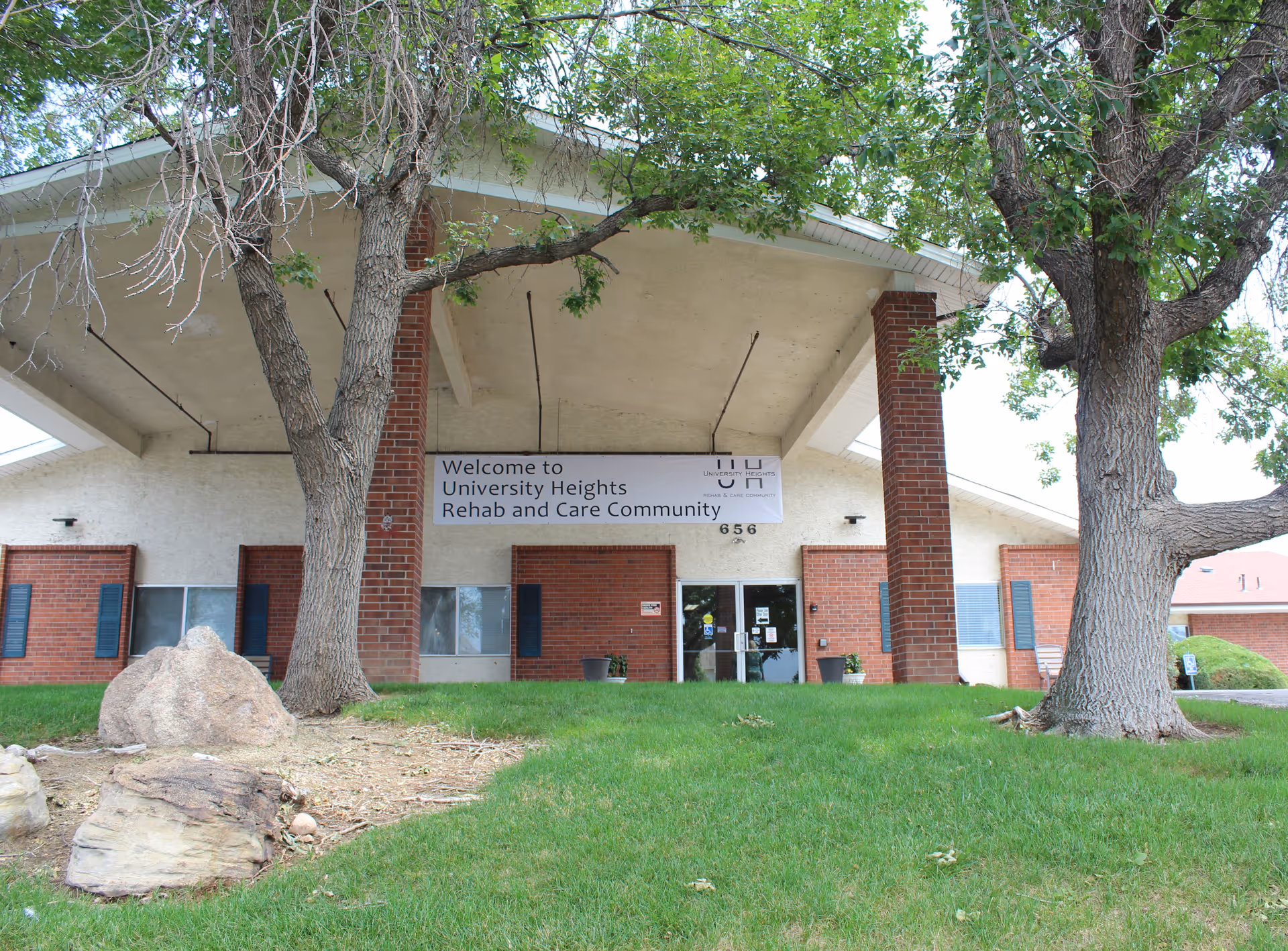 Front exterior view of University Heights Rehab and Care Community building with a covered entrance supported by brick pillars, surrounded by green grass, trees, and rocks.