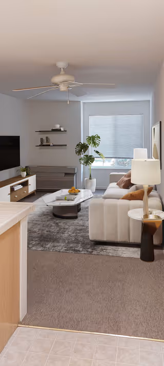 A modern living room with a beige sofa adorned with brown and orange pillows, a white marble coffee table with a bowl of oranges, a floor lamp with a white shade, a large window with closed blinds, a ceiling fan, a flat-screen TV mounted on the wall, and a potted plant near the window. The room has a gray area rug and light-colored walls.