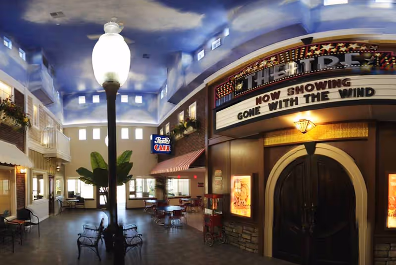 Interior view of a senior living facility designed to resemble a small town street with a sky-painted ceiling, a street lamp in the center, benches, a cafe with tables and chairs, and a theater entrance displaying a marquee that reads 'Now Showing Gone With The Wind'.