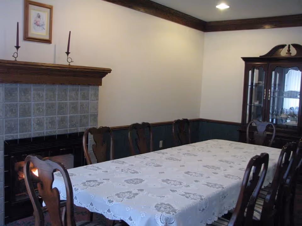 A dining room with a long table covered by a white lace tablecloth surrounded by wooden chairs. There is a fireplace with a wooden mantle and two tall candles on it. A framed picture hangs above the fireplace. In the corner, there is a wooden china cabinet with glass doors displaying dishes.