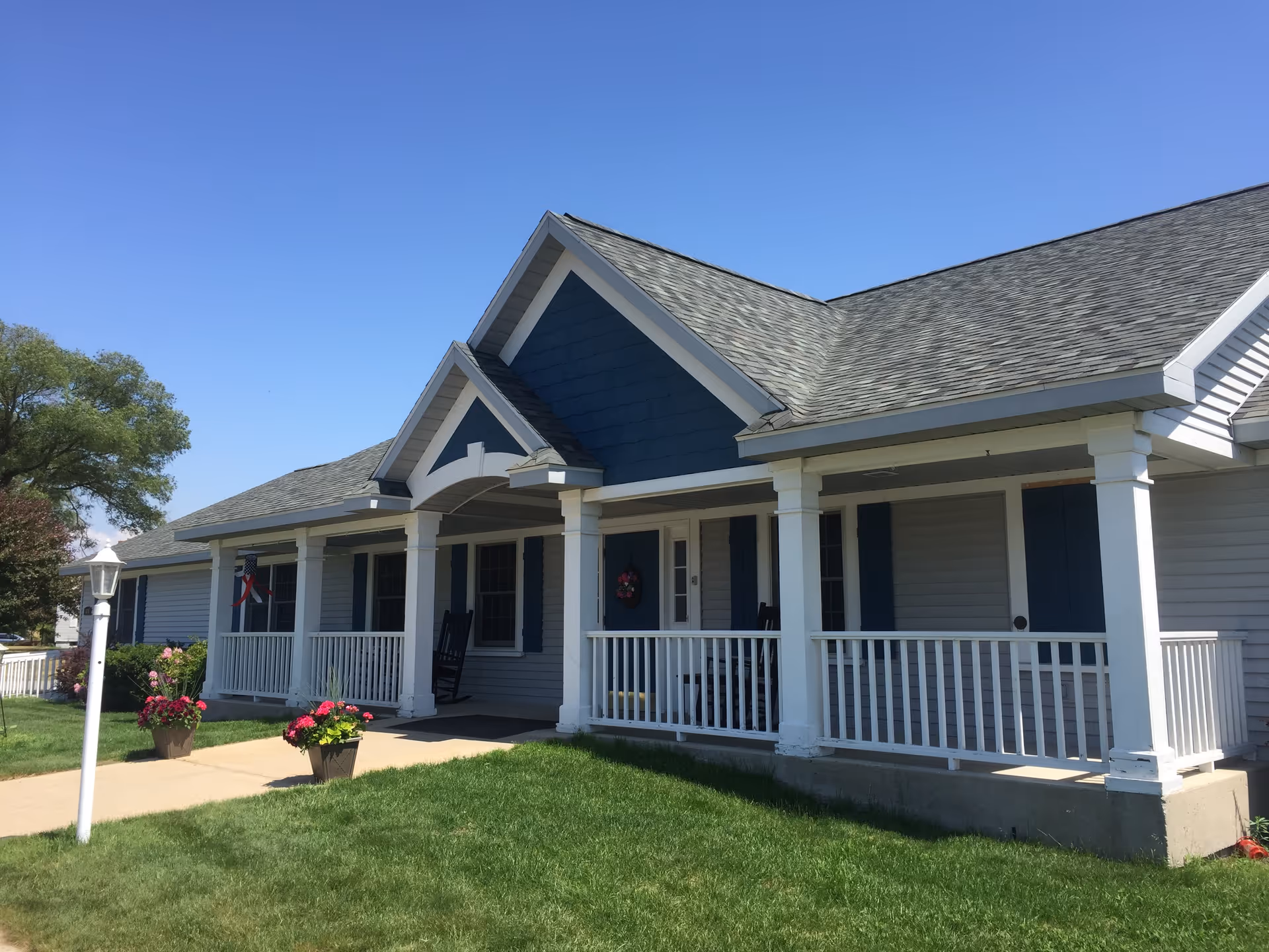 Exterior view of a single-story residential building with a gray roof, blue and gray siding, white columns, and a white railing along the front porch. There are two rocking chairs on the porch, potted flowers near the walkway, and a lamp post on the lawn. The sky is clear and blue.