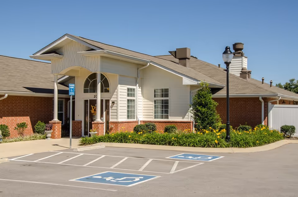 Exterior view of a senior living facility building with a covered entrance, brick and white siding walls, landscaped flower beds, and two handicap parking spaces in front under a clear blue sky.