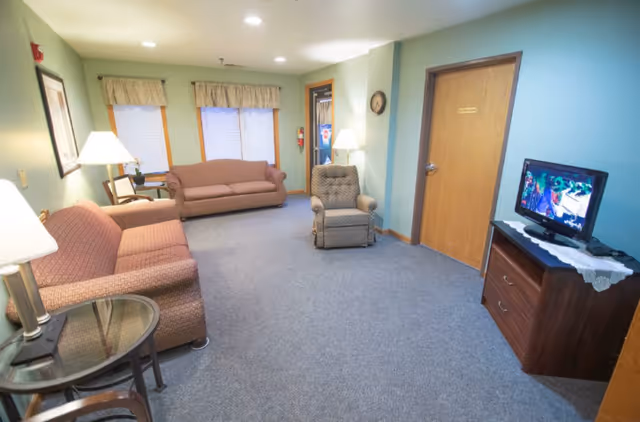A cozy living room in an assisted living facility with two upholstered sofas, a cushioned armchair, a small round glass-top table with a lamp, a wooden cabinet with a TV on top, and windows with valances letting in natural light.