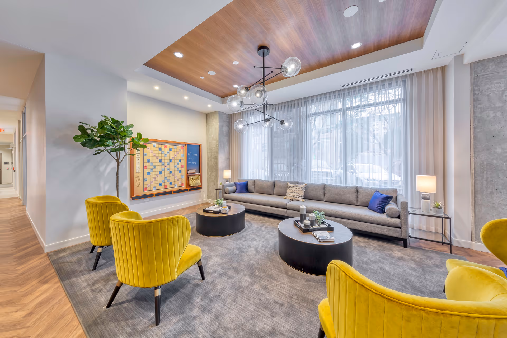 A modern living room with a long gray sofa adorned with blue and patterned pillows, four yellow velvet armchairs, two round black coffee tables with decorative items, a large window with sheer white curtains, a wooden ceiling panel with recessed lighting and a contemporary chandelier, and a wall-mounted Scrabble board with a welcome message.