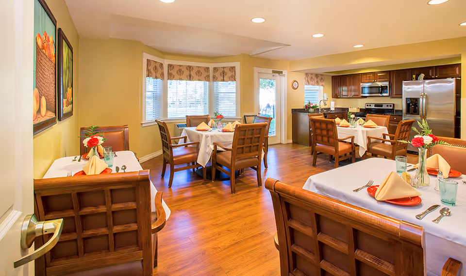 A bright dining room with wooden floors and multiple tables set with white tablecloths, folded napkins, plates, and glassware. The room features wooden chairs with cushioned seats, a bay window with patterned valances, and a kitchen area with stainless steel appliances including a refrigerator, microwave, and stove. There are framed fruit paintings on the wall and floral centerpieces on the tables.