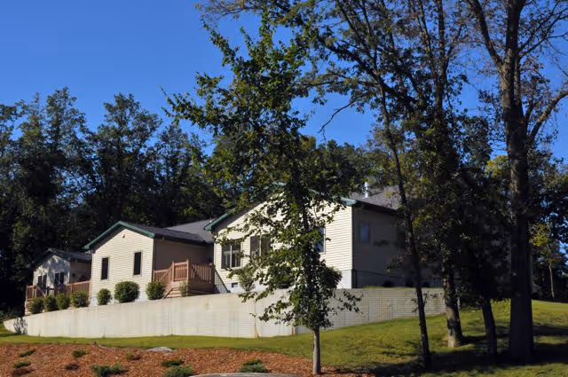Exterior view of a single-story residential building with beige siding and a dark roof, surrounded by trees and a grassy area with a retaining wall in front.