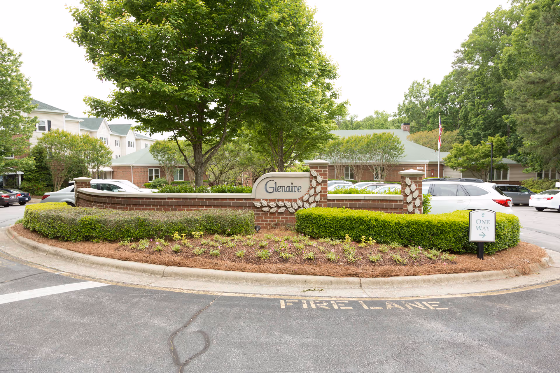 Entrance area of the Glenaire facility featuring a brick sign with the name 'Glenaire' surrounded by landscaped bushes and trees. Several cars are parked in the background near buildings with green roofs. A small sign indicating 'One Way' is visible on the right side.