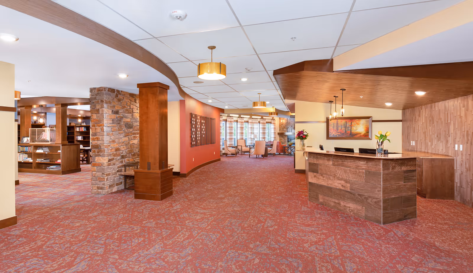 Spacious interior of a senior living facility with a large wooden reception desk on the right, a stone pillar and wooden shelves on the left, and a seating area with chairs and a fireplace in the background. The space features warm lighting, red patterned carpet, and wood accents on the ceiling and walls.