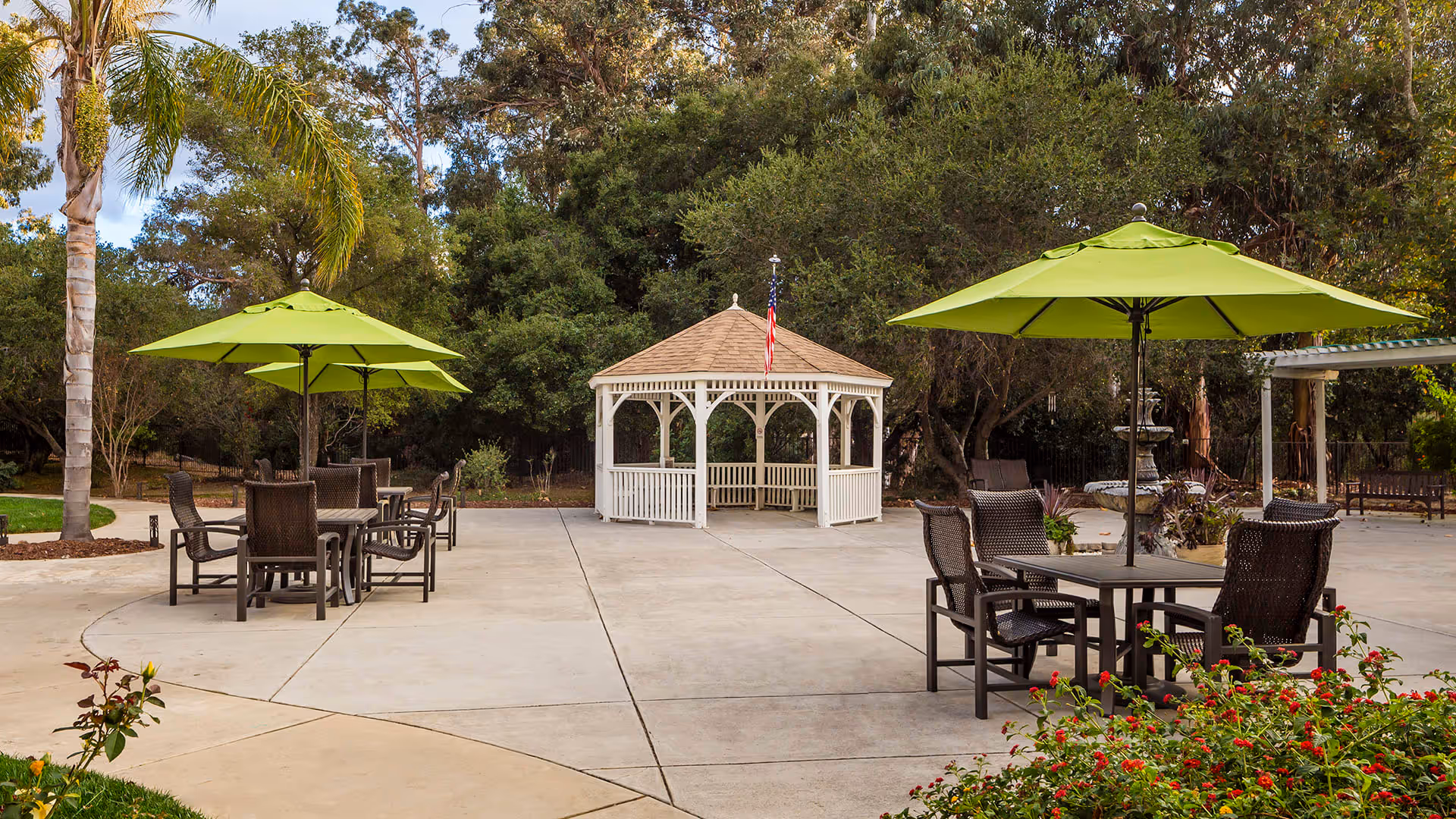 Outdoor patio area with several tables and chairs under green umbrellas, surrounded by trees and plants. In the background, there is a white gazebo with an American flag on top.
