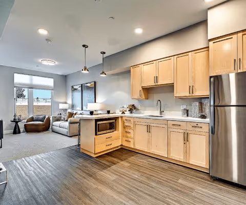 Open-concept senior living suite with a kitchenette featuring light wood cabinets and a stainless-steel fridge beside a living area with a sofa and large window.