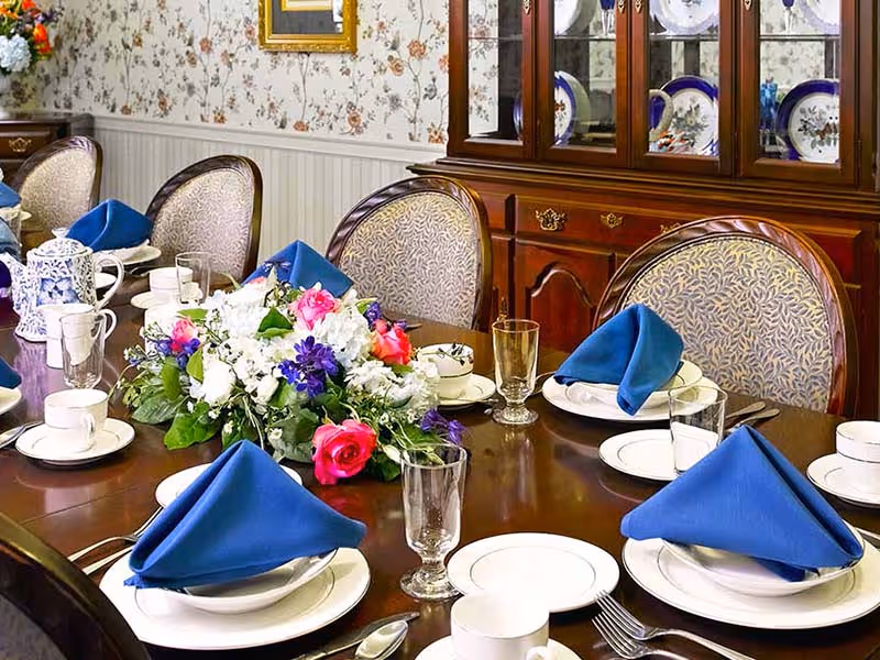 A formal dining room table set with white plates, cups, and glasses, each place setting featuring a neatly folded blue napkin. A floral centerpiece with white, pink, and purple flowers decorates the middle of the polished wooden table. Upholstered chairs with patterned fabric surround the table, and a wooden china cabinet with displayed plates is visible in the background against floral wallpaper.