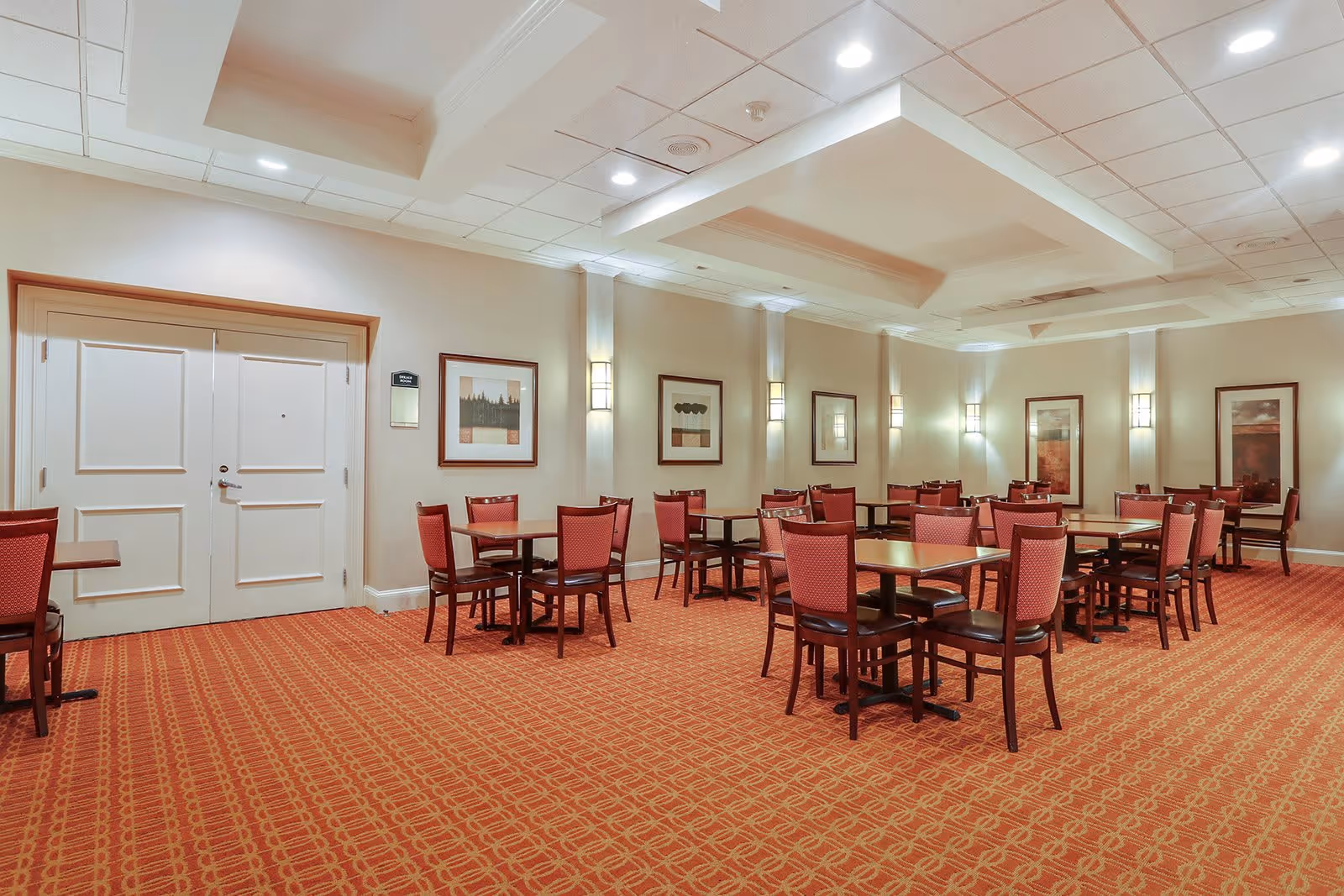 A dining room with multiple square wooden tables and red cushioned chairs arranged neatly on a patterned red carpet. The walls are beige with framed artwork and wall-mounted lights. The ceiling has recessed lighting and decorative beams. There is a double door on one side of the room.