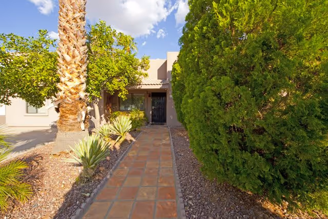 Tiled walkway through desert landscaping and trees leading to the front entrance of a single-story building.