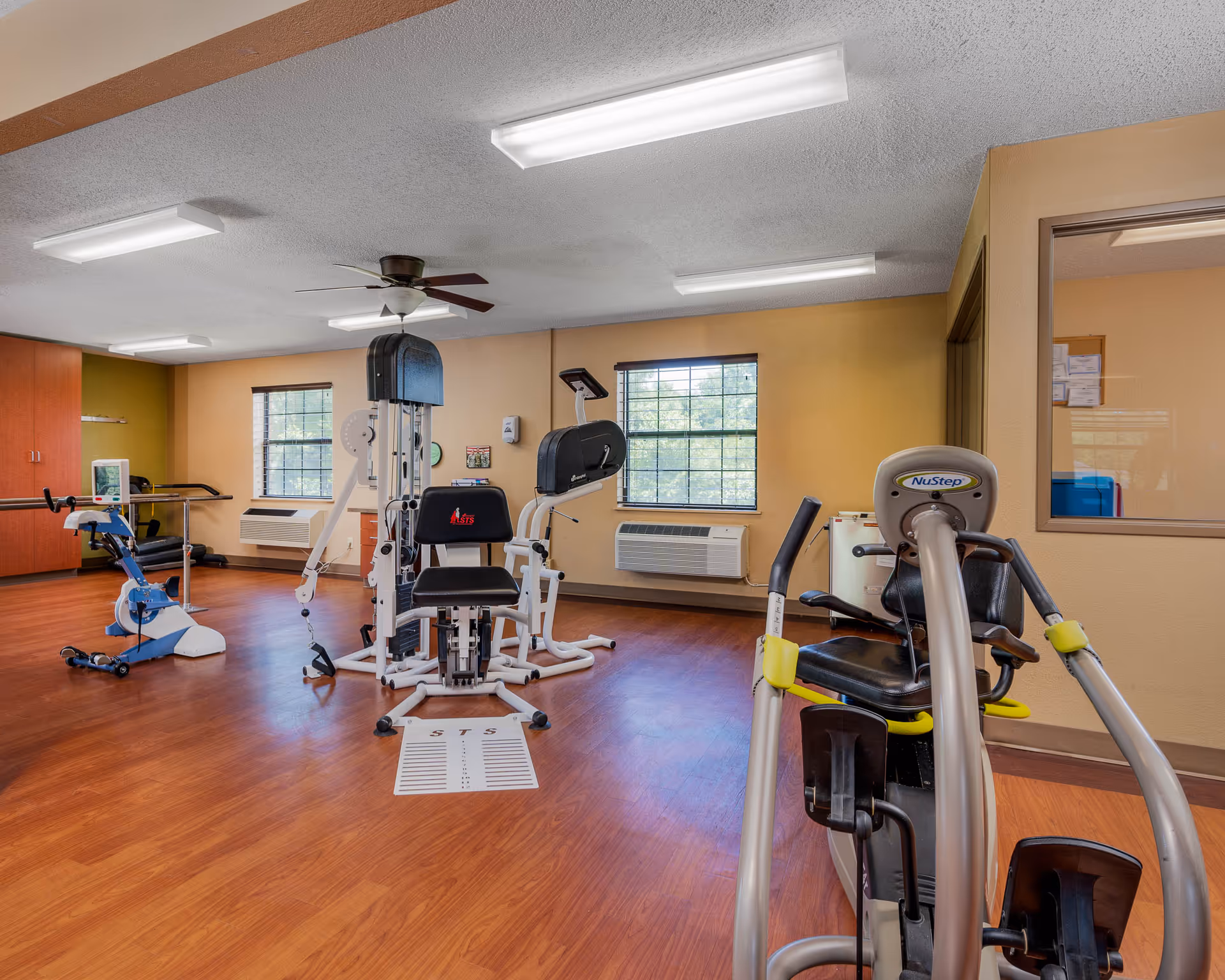 A rehabilitation and exercise room with various fitness equipment including a stationary bike, a multi-function exercise machine, and a NuStep recumbent cross trainer. The room has wood flooring, beige walls, two windows with black frames, ceiling lights, and a ceiling fan. There is a windowed office or room visible on the right side.