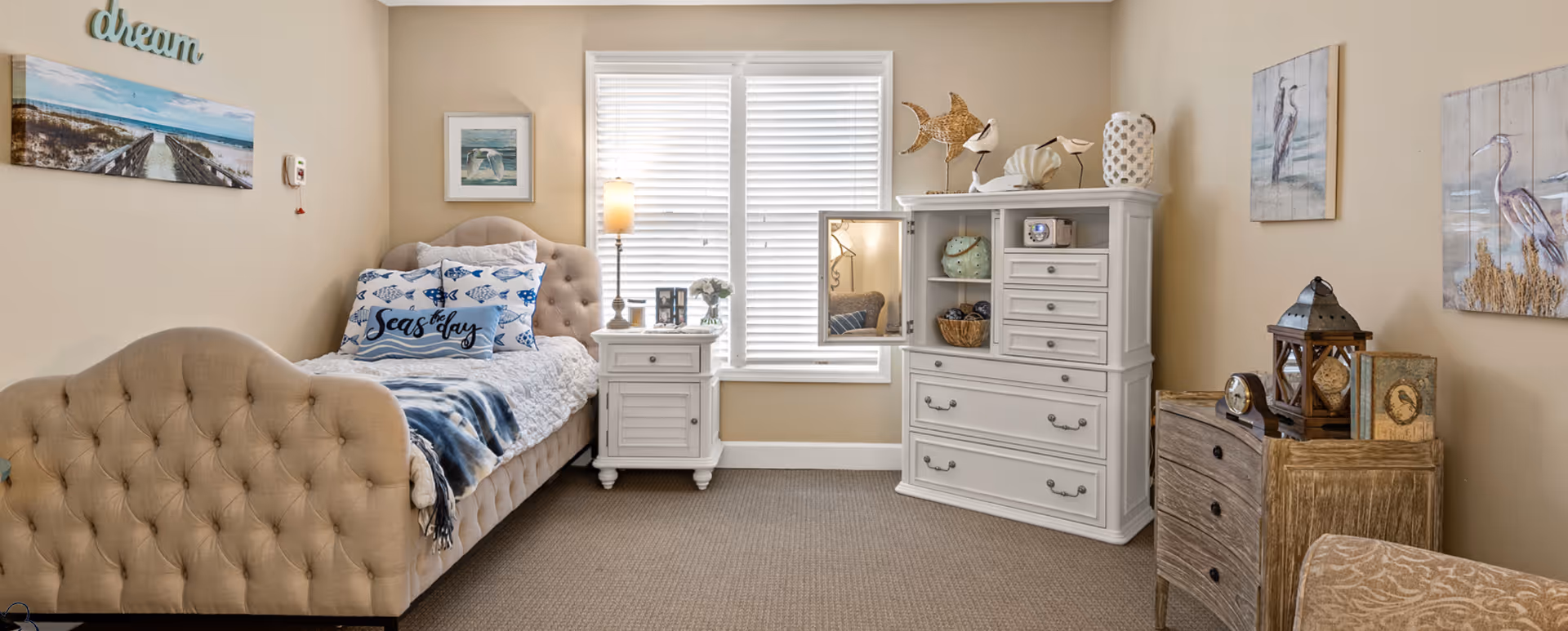 A cozy bedroom with a beige tufted bed adorned with blue and white fish-themed pillows and a blanket. Next to the bed is a white nightstand with a lamp, picture frames, and a small flower vase. A white dresser with drawers and shelves holds decorative items including a woven fish sculpture and bird figurines. The walls are decorated with coastal-themed artwork and the window has white blinds. The room has a warm and inviting atmosphere.