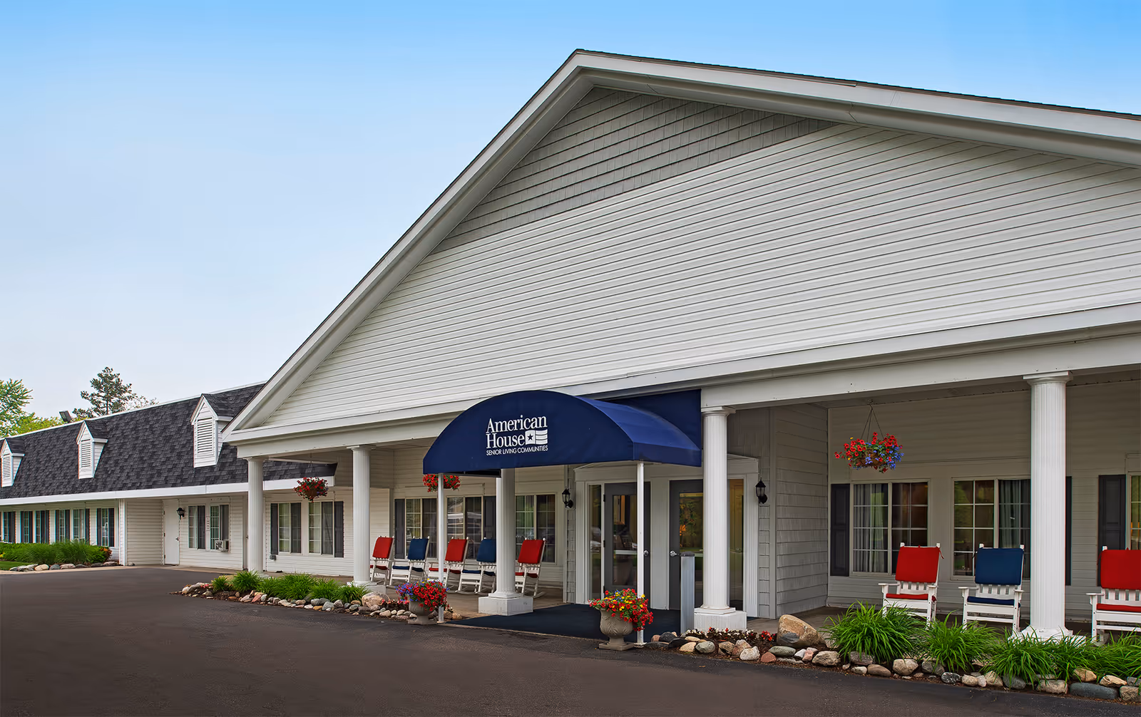 Exterior view of American House Dearborn Heights senior living community building with white siding, a blue awning over the entrance, red and blue rocking chairs on the porch, hanging flower baskets, and landscaping with rocks and greenery.