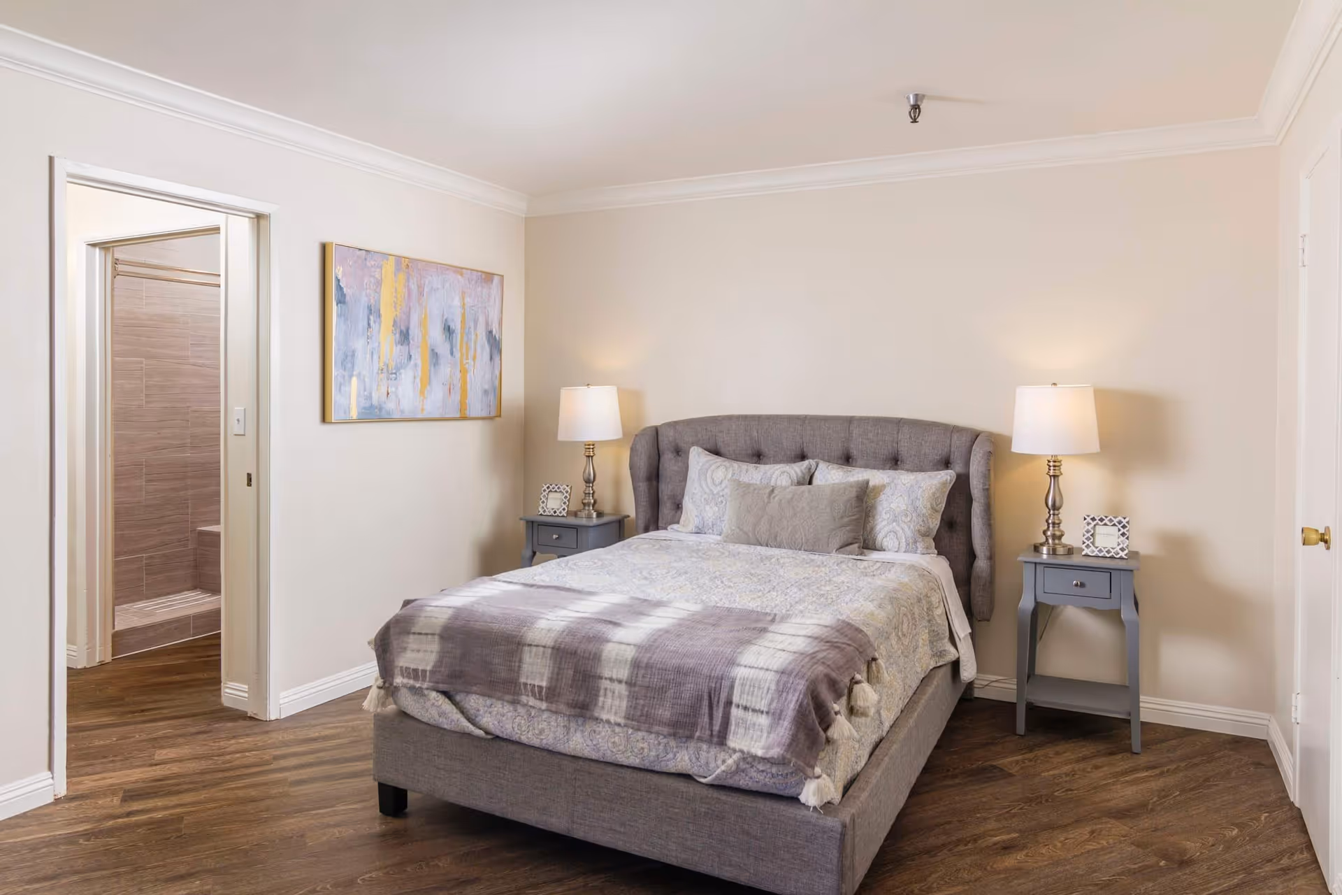 A neatly made bed with a tufted gray headboard in a bedroom. The bed has multiple pillows and a patterned blanket with a plaid throw at the foot. There are two matching gray nightstands on either side of the bed, each with a lamp and a small decorative item. A doorway on the left leads to a bathroom with a tiled shower. The walls are painted light beige, and there is a colorful abstract painting hanging on the wall to the left of the bed.