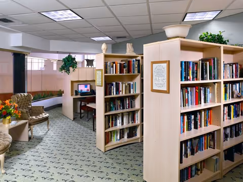 Interior view of a library or reading area in a senior living facility with bookshelves filled with books, a computer workstation, comfortable chairs, potted plants, and soft lighting.