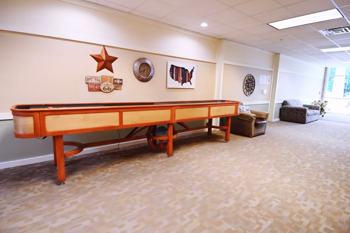 A long wooden shuffleboard table in a bright senior living lounge with chairs, sofas, and wall decorations.