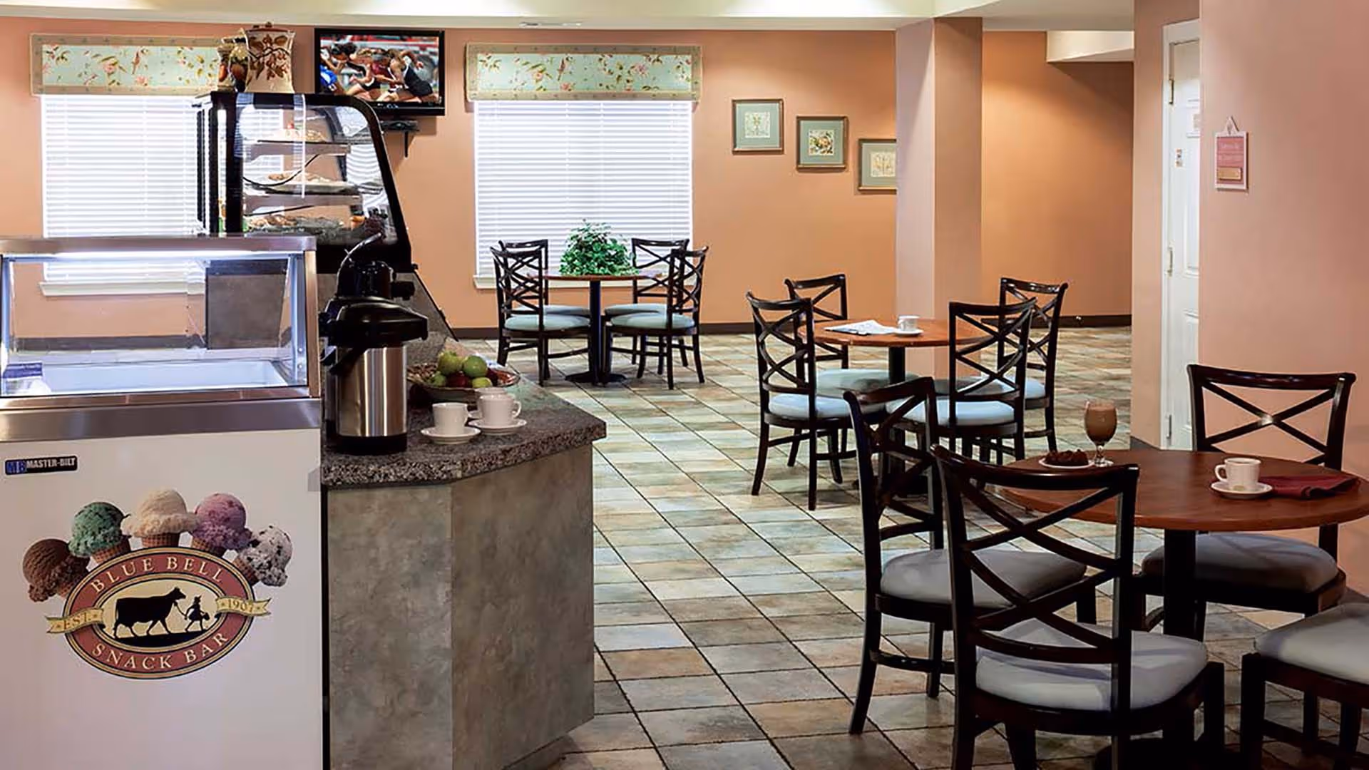 A cozy dining area with several round wooden tables and cushioned chairs arranged on a tiled floor. There is a snack bar counter with a coffee dispenser and cups, and an ice cream freezer with a Blue Bell Snack Bar logo. The walls are painted a warm peach color, decorated with framed artwork and windows with floral valances.