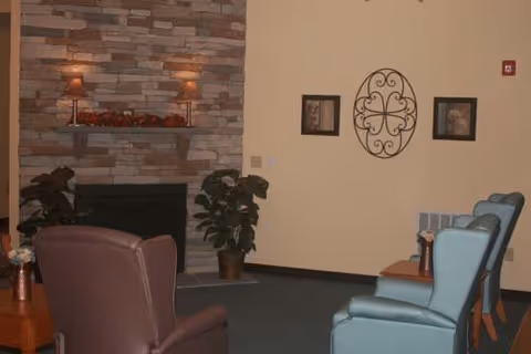 Comfortable seating area with armchairs facing a stone fireplace and wall decor in a senior living facility common room.