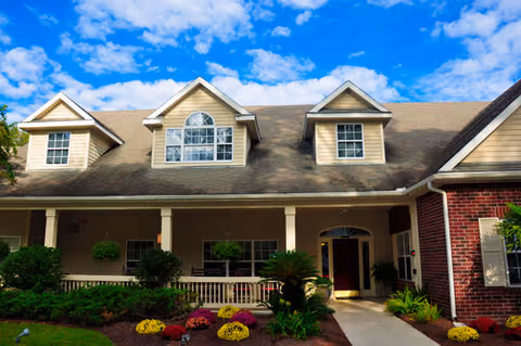 Front exterior view of a residential-style assisted living facility with beige siding and brick accents, featuring a covered porch with columns, multiple windows including dormer windows, landscaped garden with colorful flowers, shrubs, and a clear blue sky with some clouds.