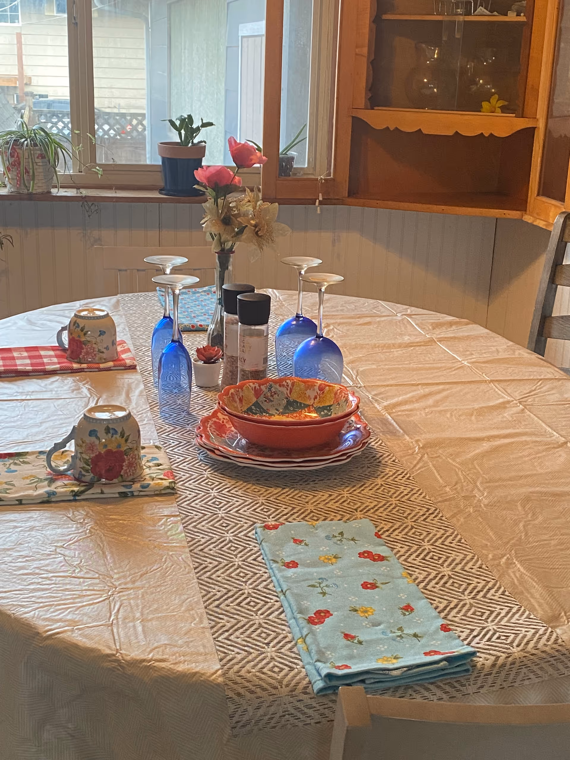 A dining table set with a beige tablecloth and a patterned table runner. On the table are four blue wine glasses turned upside down, two floral mugs on colorful napkins, a stack of colorful plates with a bowl on top, salt and pepper shakers, and a small vase with artificial flowers. In the background, there is a window with potted plants on the sill and a wooden cabinet with glass doors.