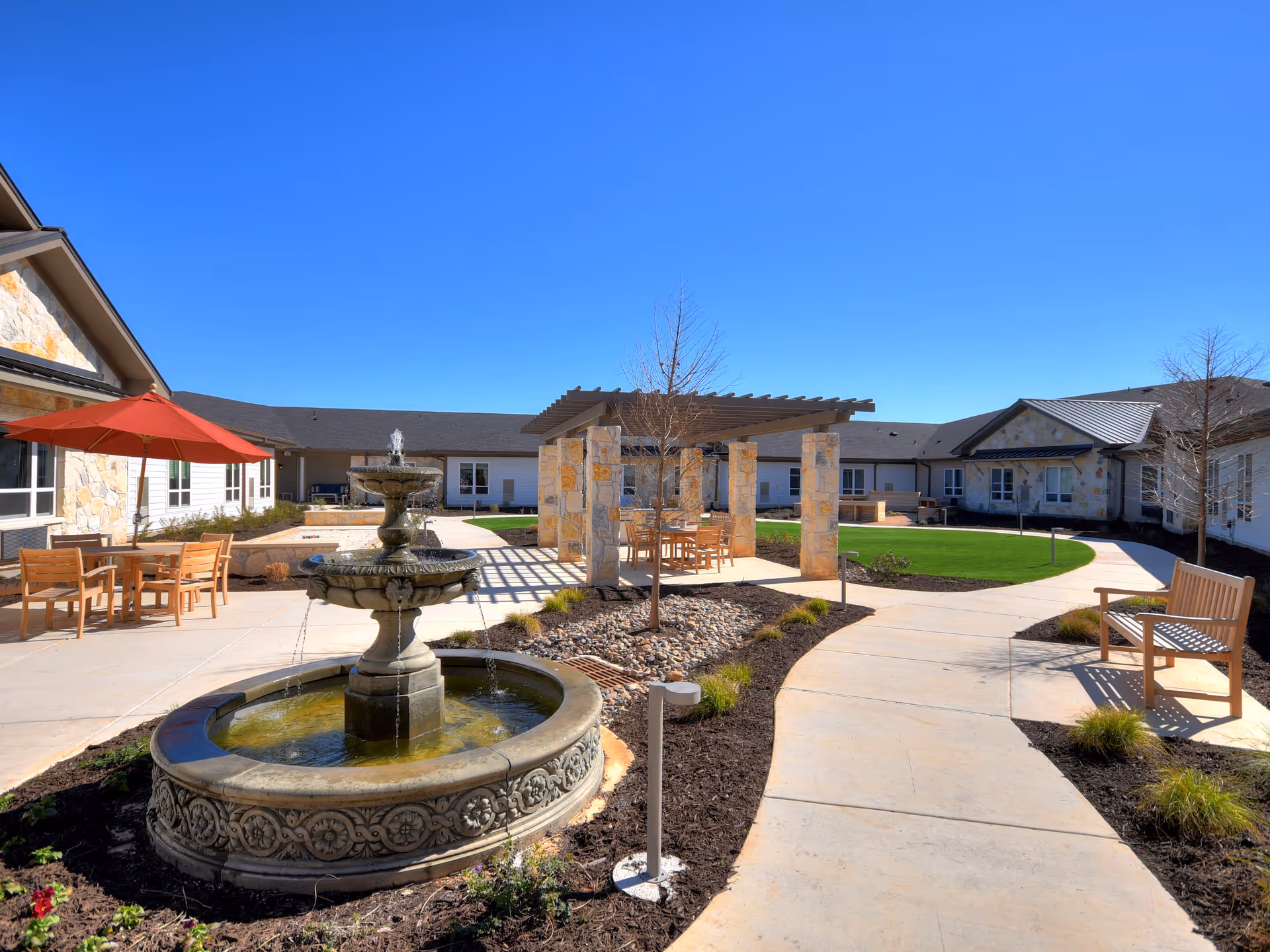 Outdoor courtyard area of a senior living facility featuring a stone water fountain in the foreground, wooden benches, tables with chairs under a red umbrella and a pergola, surrounded by a paved walkway, grass, and buildings under a clear blue sky.