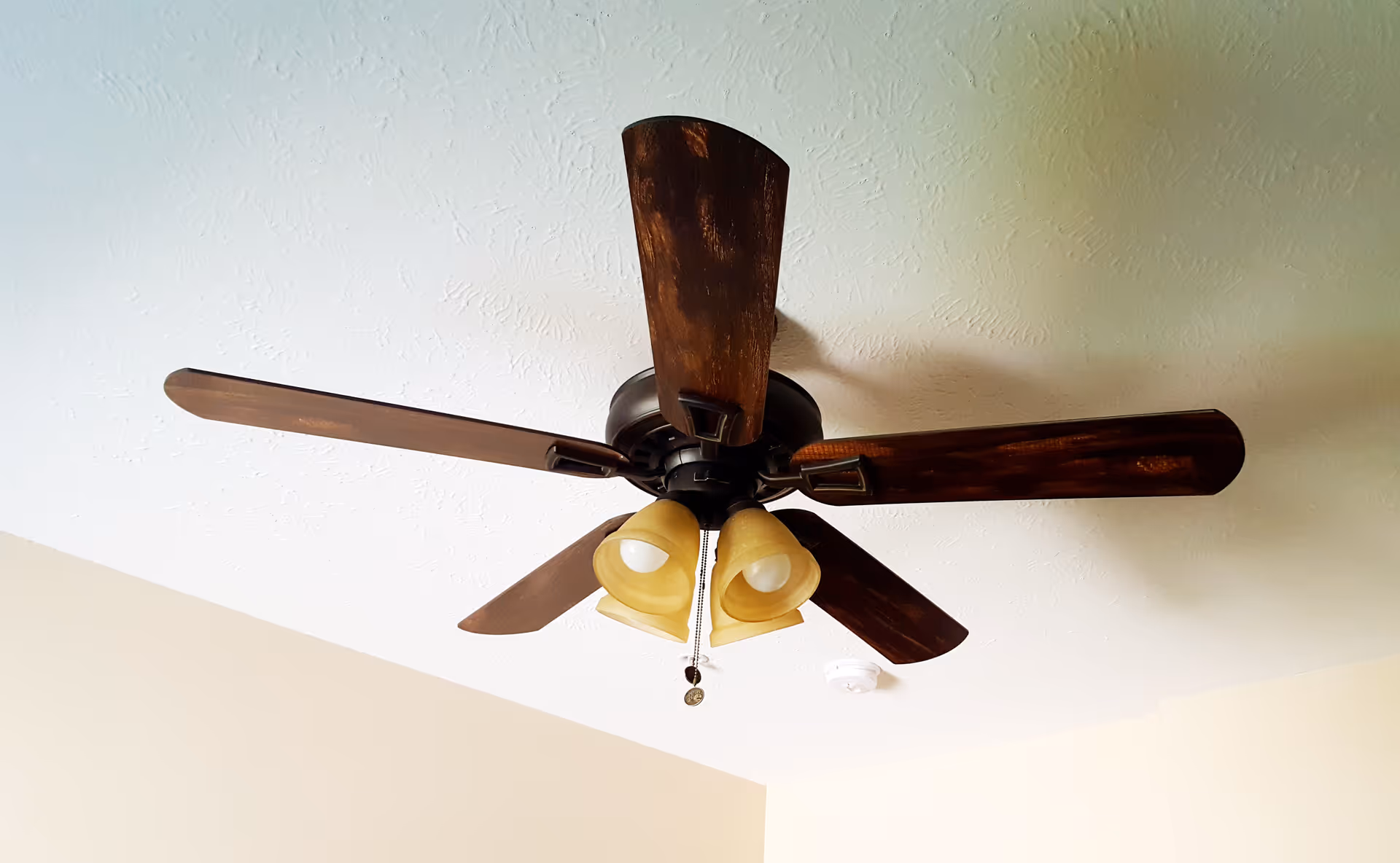 Ceiling fan with five dark wooden blades and three downward-facing light fixtures with yellowish glass shades mounted on a white textured ceiling.