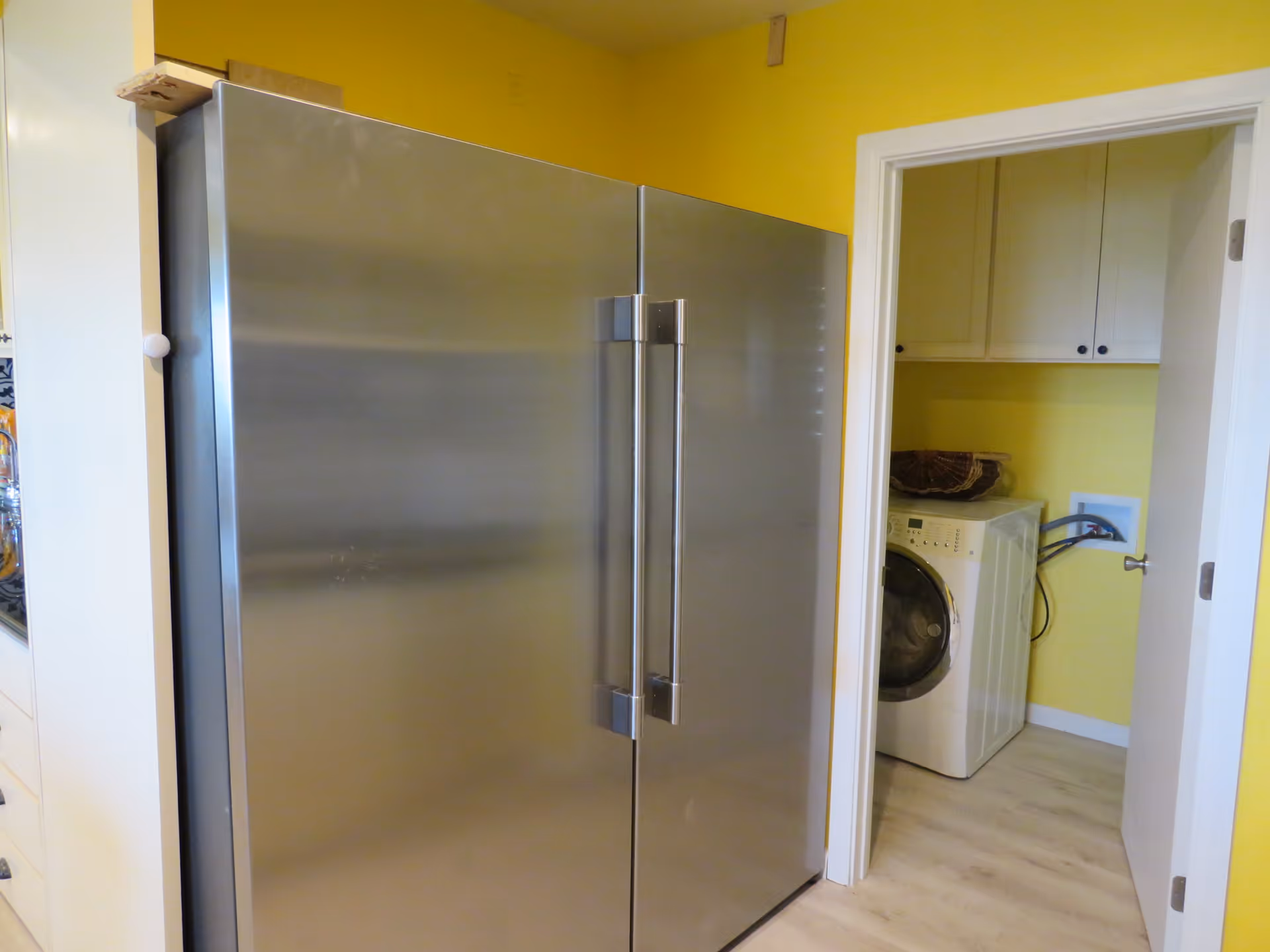 A modern kitchen area with a large stainless steel double-door refrigerator on the left and an adjacent laundry room visible through an open door, featuring a front-loading washing machine and white cabinets against yellow walls.